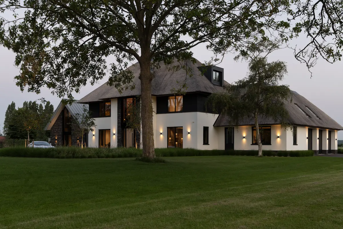 Moderne villa met rieten kap, witte gevels en zwarte accenten, sfeervol verlicht en omringd door een ruime groene tuin met volwassen bomen.