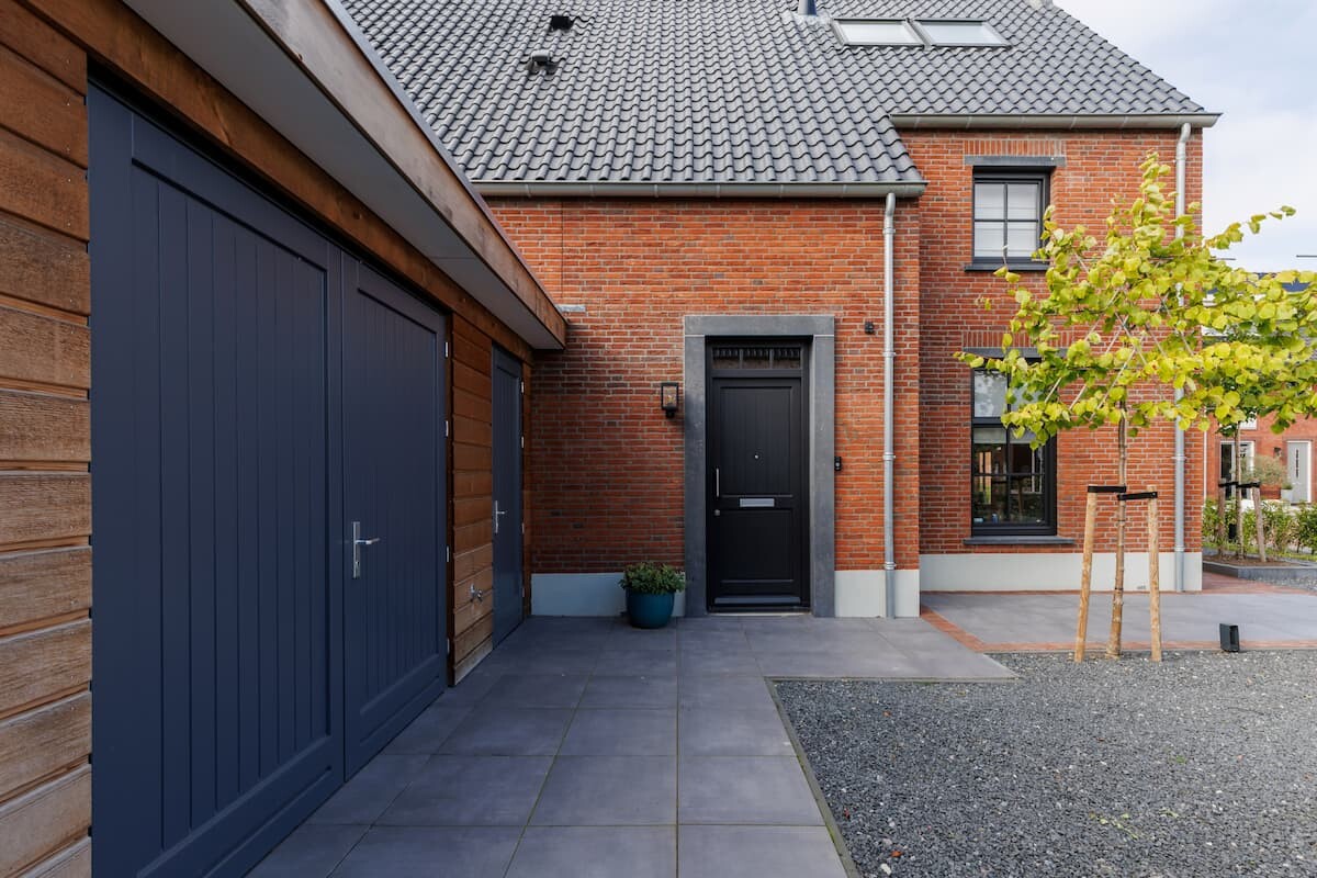 De ingang van een woning met een bakstenen gevel, een houten schuurdeurtje, een moderne zwarte voordeur en een goed onderhouden oprit met tegels en grind.