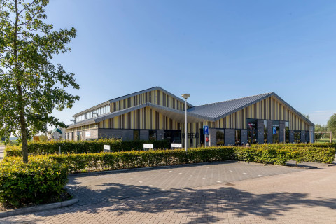 Multifunctioneel gebouw MFA De Beemd in Oud-Alblas, met een modern ontwerp en een gevel van verticale houten en metalen panelen. Het pand heeft een speels dakontwerp en wordt omringd door groen en een ruim parkeerterrein.