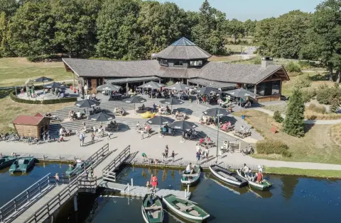 Houten paviljoen met groot terras aan het water, vol bezoekers en omringd door bos en roeiboten aan een steiger.