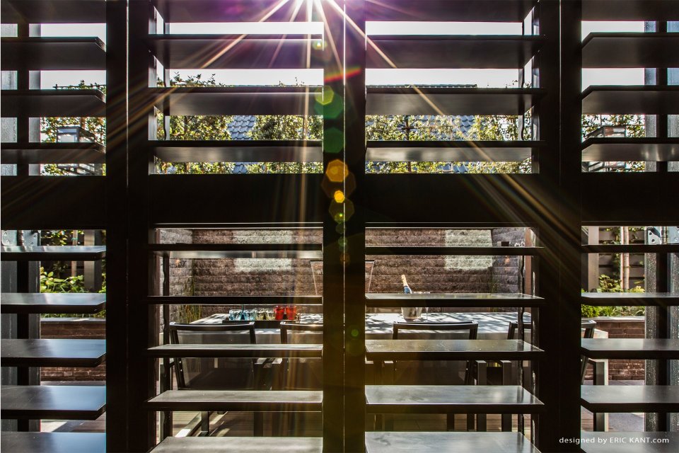 Zonnestralen door houten shutters met zicht op terras &ndash; licht en zicht in architectuur als sfeerbepalend element in het ontwerp.
