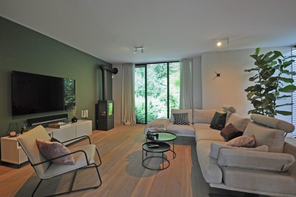 Lichte woonkamer met houten vloer, hoekbank en houtkachel, met zicht op groene tuin &ndash; modern en sfeervol design door Bongers Architecten