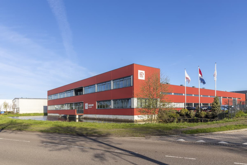 "Modern rood bedrijfsgebouw met grote raampartijen en strakke belijning, gelegen aan een brede weg met groenstroken. Voor het pand wapperen drie vlaggen, waaronder de Nederlandse vlag."